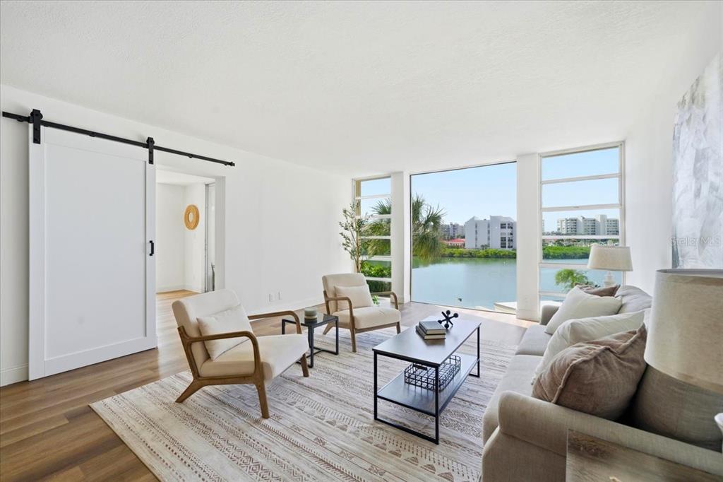 A bright living room features a sliding barn door, neutral furniture, and large waterfront windows — 7400 SUN ISLAND DRIVE S UNIT 202, SOUTH PASADENA, FL