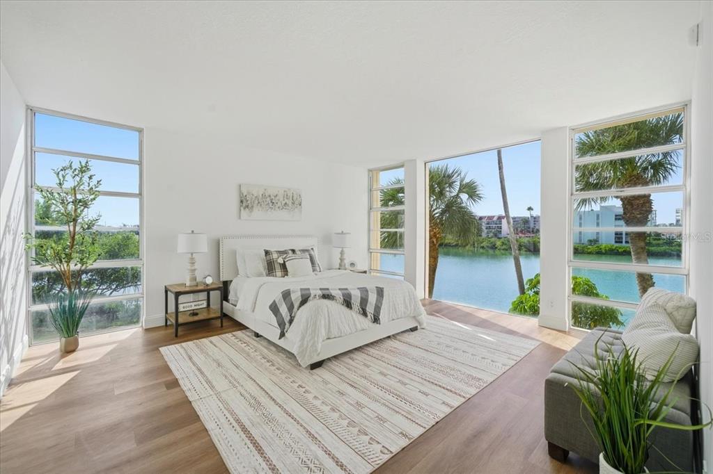 A bright bedroom features a bed, large windows overlooking water, and light wood flooring — 7400 SUN ISLAND DRIVE S UNIT 202, SOUTH PASADENA, FL