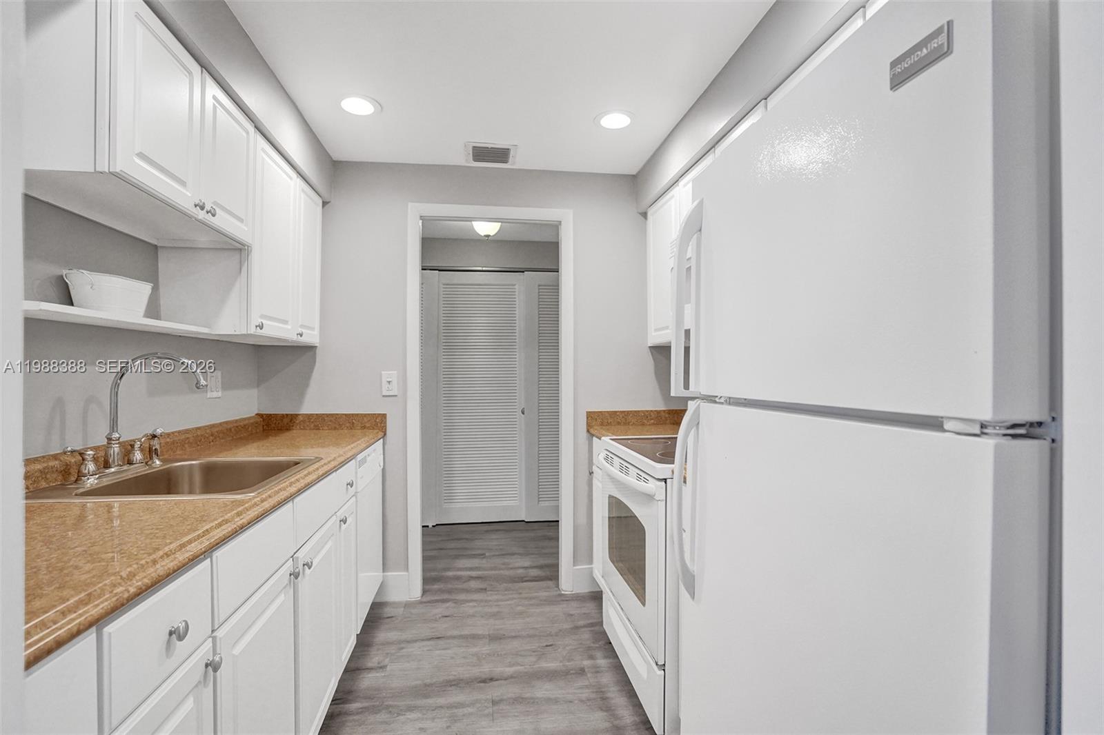 A galley kitchen features white cabinets, tan countertops, white appliances, and gray wood-look flooring — 400 N Riverside Dr # 214, Pompano Beach FL 33062, POMPANO BEACH, FL
