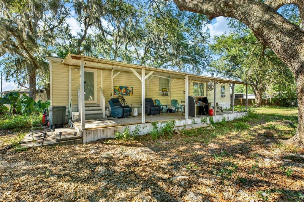 A light-yellow mobile home features a long, covered wooden porch shaded by large oak trees — 3642 KALMAN DRIVE, ZEPHYRHILLS, FL