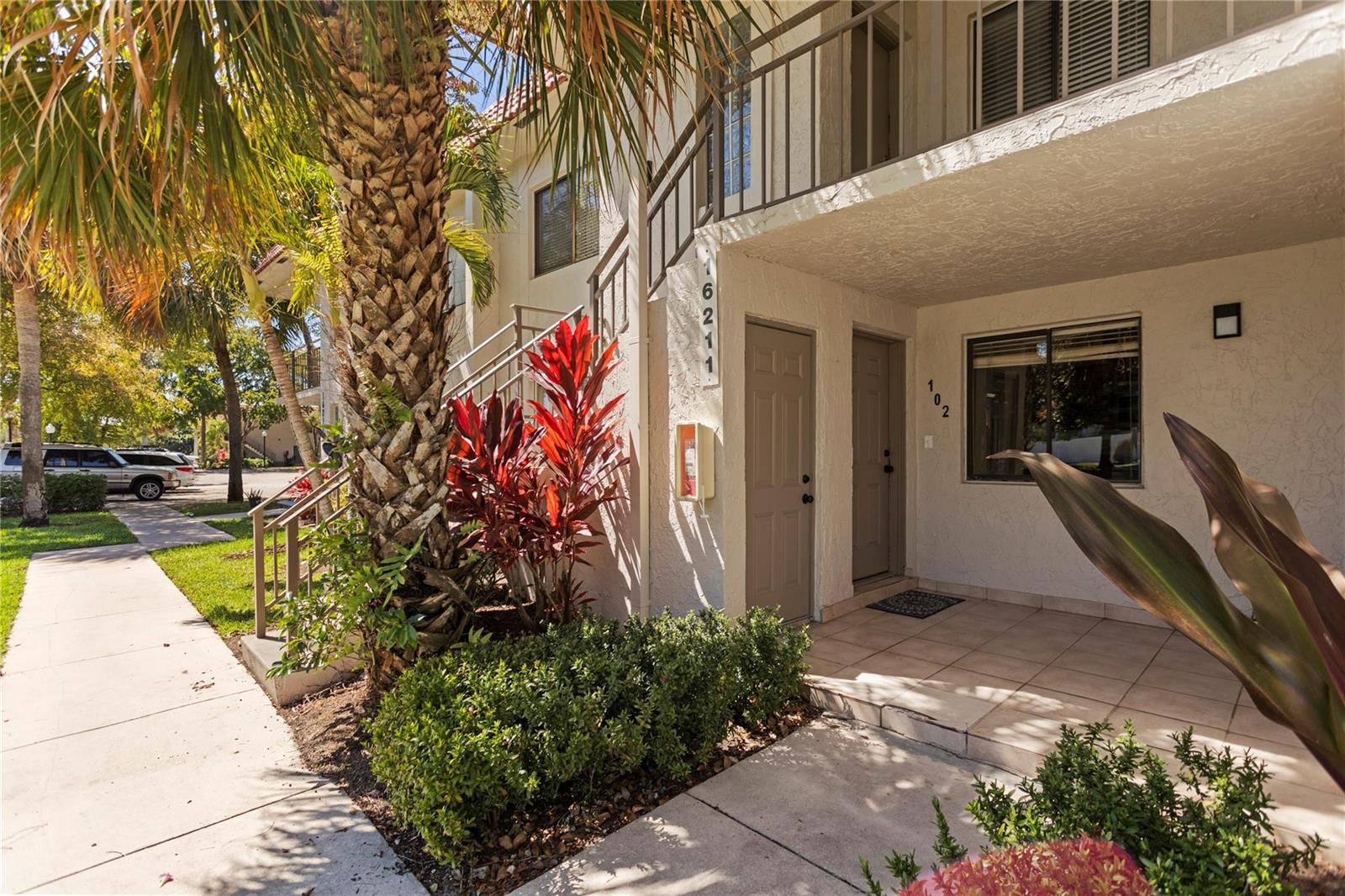 A ground-level apartment entrance with a tiled patio, beige stucco walls, and tropical landscaping — 16211 Saddle Club Road # 102, Weston FL 33326, WESTON, FL
