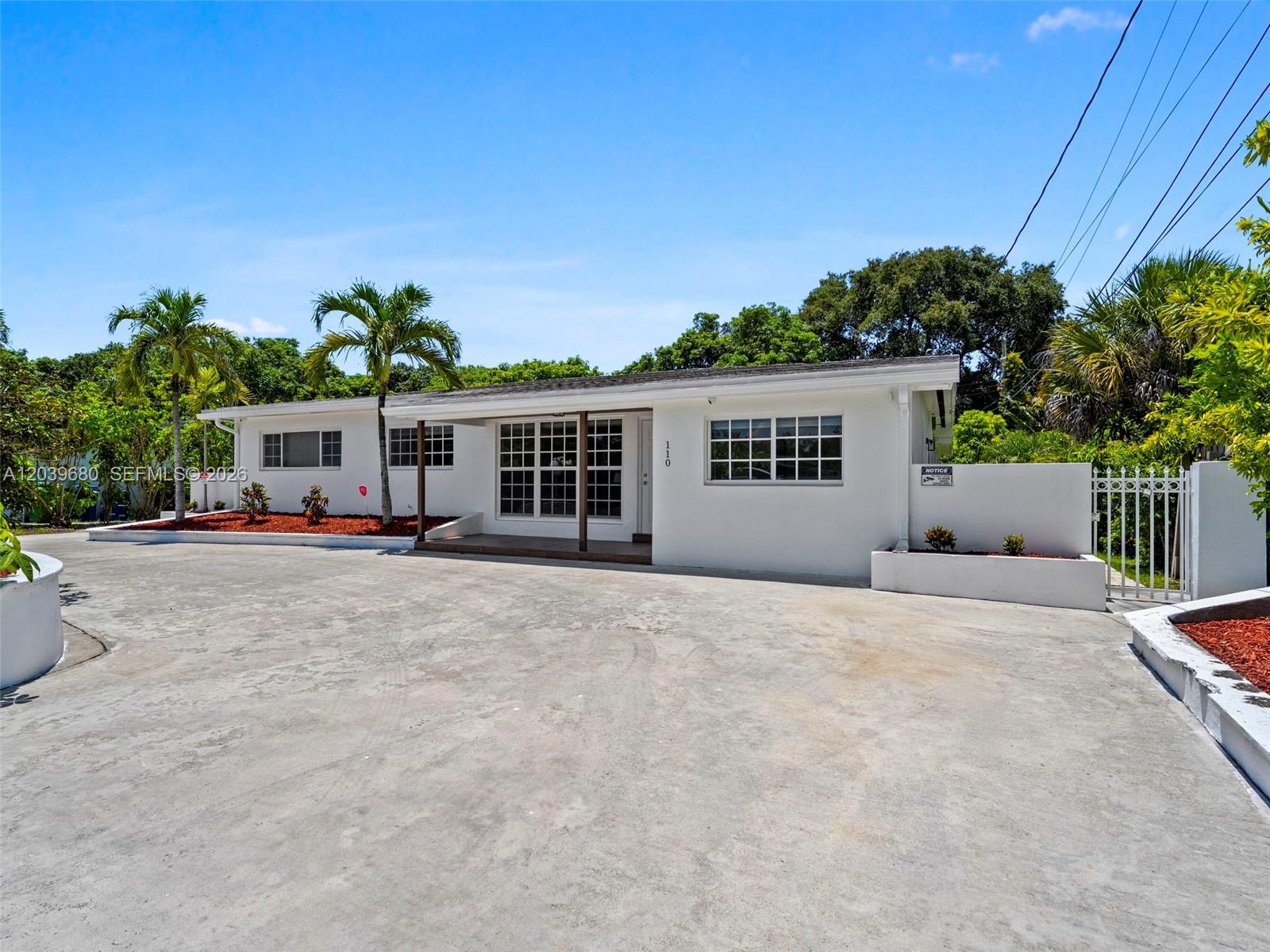 A white, single-story house features a large concrete driveway, palm trees, and a gated entrance — 110 NE 152nd St, Miami FL 33162, MIAMI, FL
