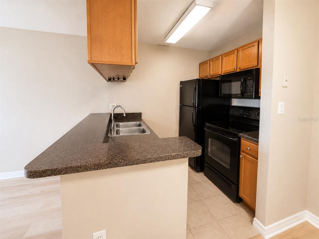A kitchen features dark countertops, black appliances, and wooden cabinets over a tiled floor — 203 LAKE PARSONS GREEN UNIT 108, BRANDON, FL
