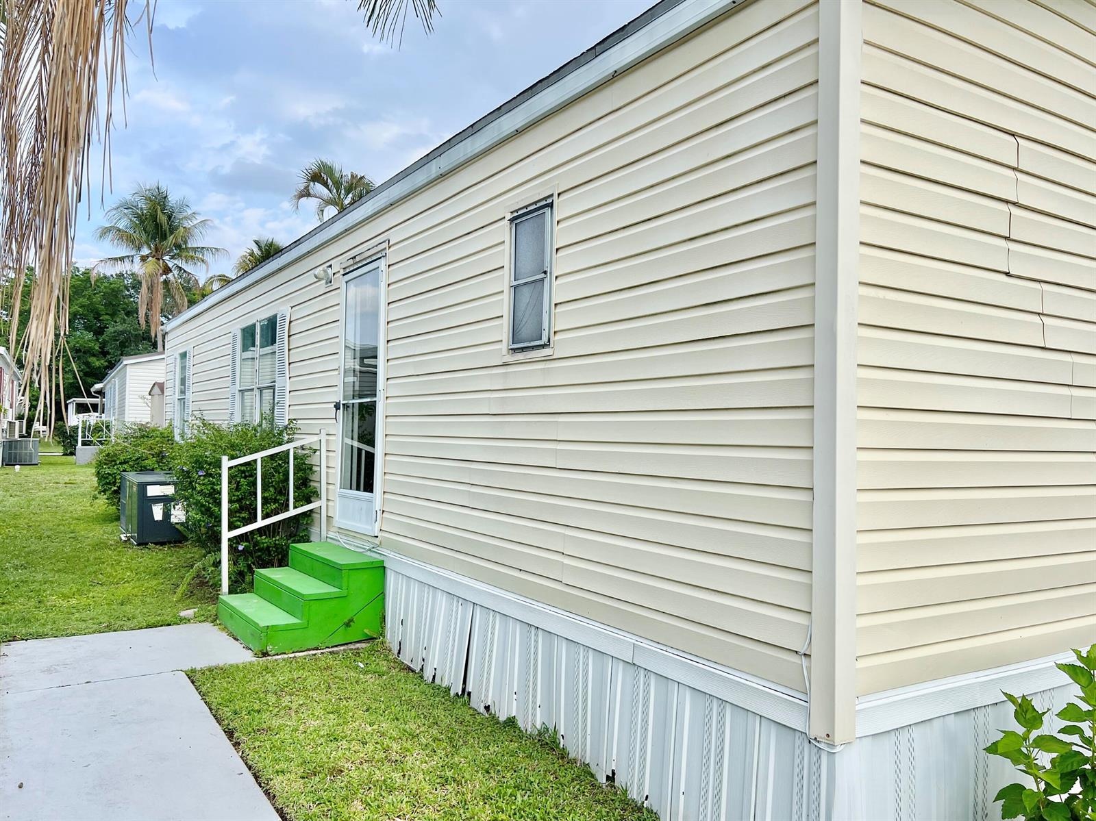 The exterior of a beige mobile home features green steps, white skirting, and a window — 183 Plantation Boulevard, Lake Worth FL 33467, LAKE WORTH, FL