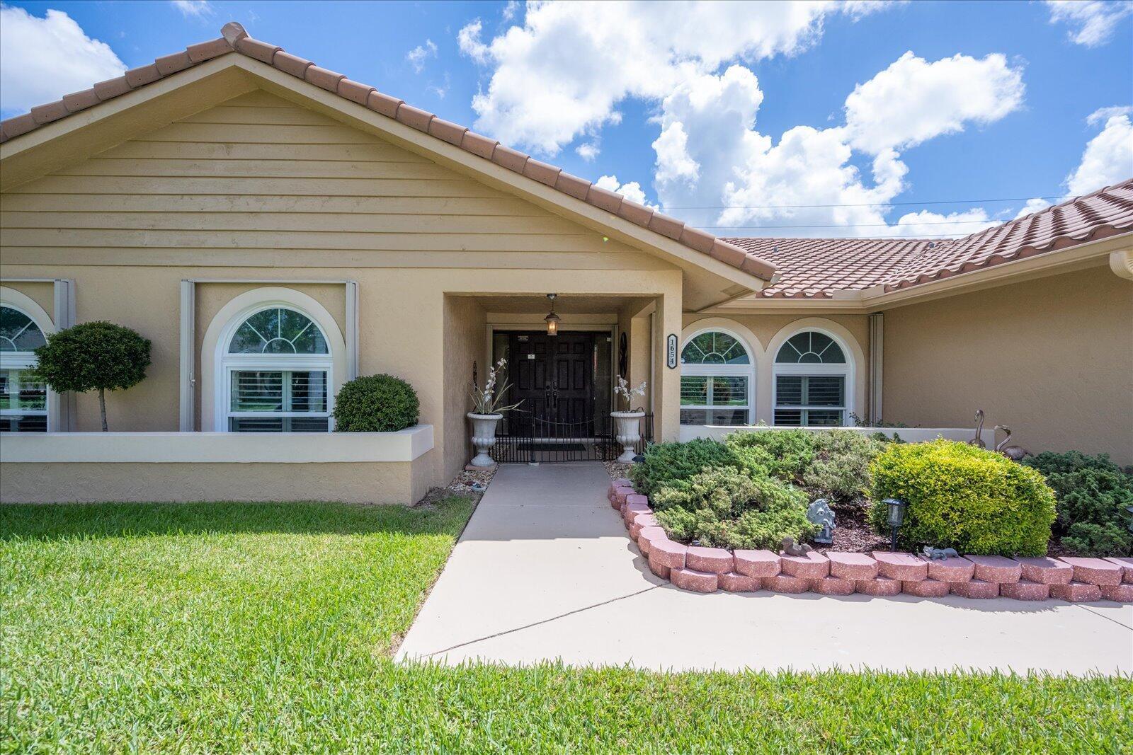 The exterior of a tan house features a covered entryway, arched windows, and landscaping — 1654 Farmington Circle, Wellington FL 33414, WELLINGTON, FL