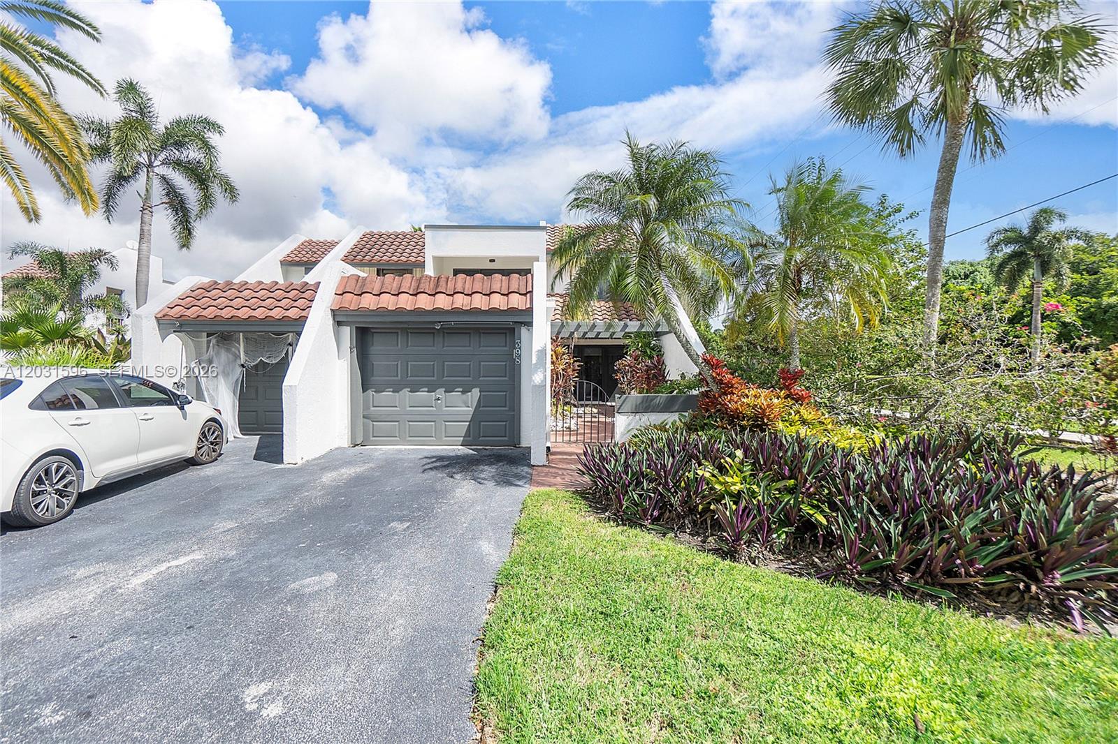 A white two-story townhouse features a gray garage, a tiled roof, and tropical landscaping — 398 Fern Dr # 6, Weston FL 33326, WESTON, FL
