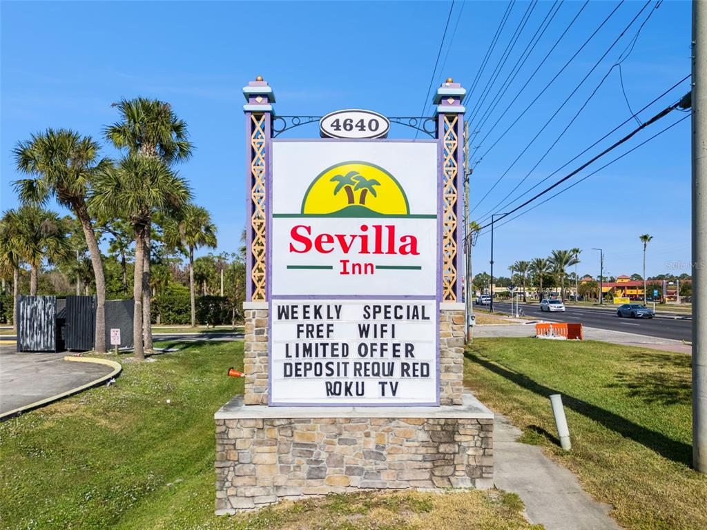 A roadside sign for the Sevilla Inn stands on a stone base near palm trees — 4640 W IRLO BRONSON MEMORIAL HIGHWAY, KISSIMMEE, FL