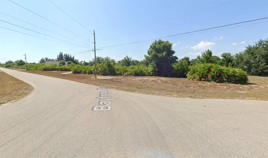 This exterior view shows a paved road intersection next to a vacant, brush-covered lot — 320 BAYTREE DRIVE, ROTONDA WEST, FL