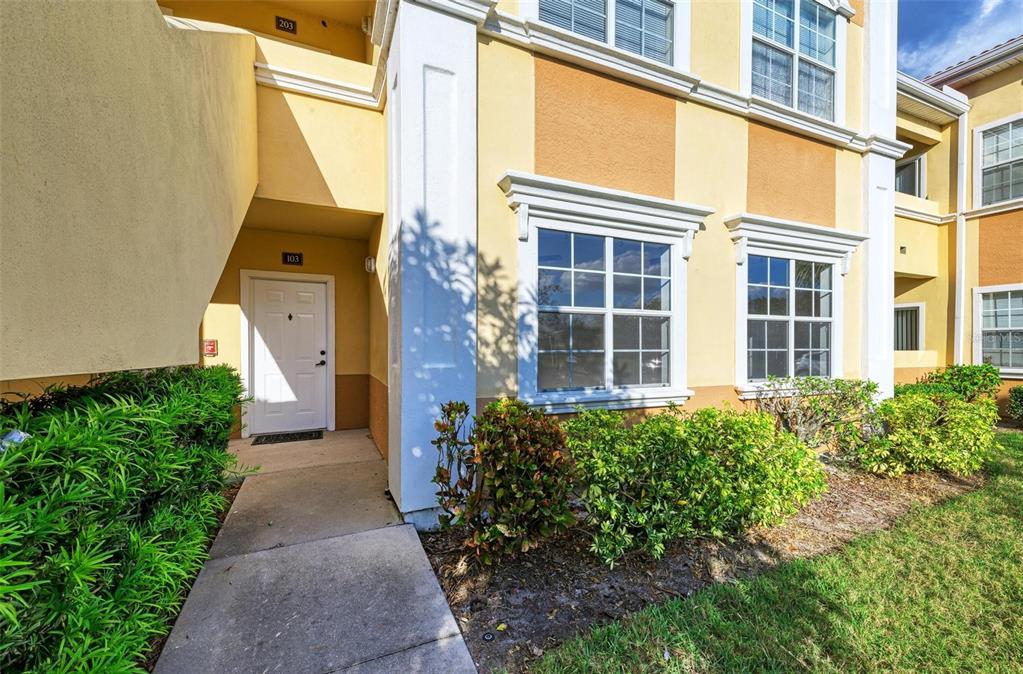 A white front door of a yellow apartment building is surrounded by green landscaping — 3005 CHIANTI COURT UNIT 103, SARASOTA, FL