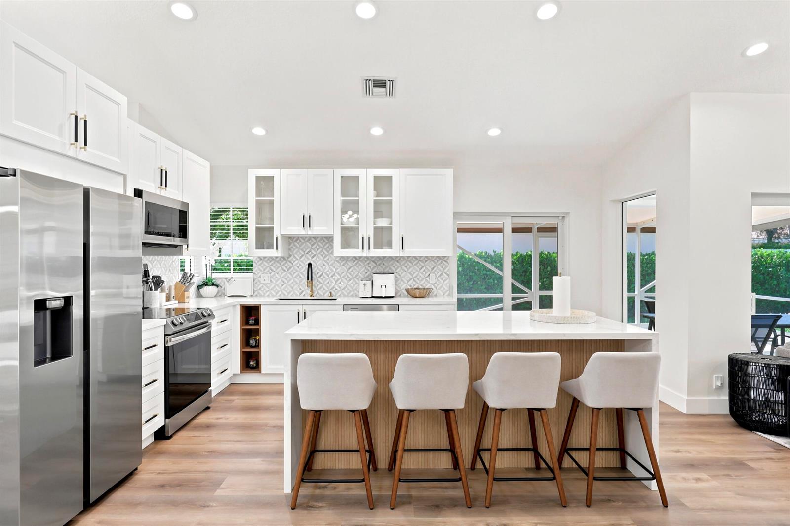 A modern white kitchen features a large island with four stools and wood-toned paneling — 5212 Bodega Place, Delray Beach FL 33484, DELRAY BEACH, FL