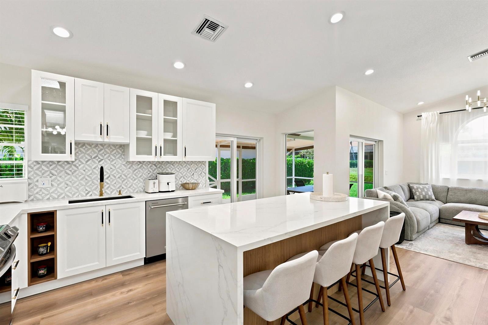 A modern kitchen with white cabinets, a marble island, and bar stools overlooks a living area — 5212 Bodega Place, Delray Beach FL 33484, DELRAY BEACH, FL