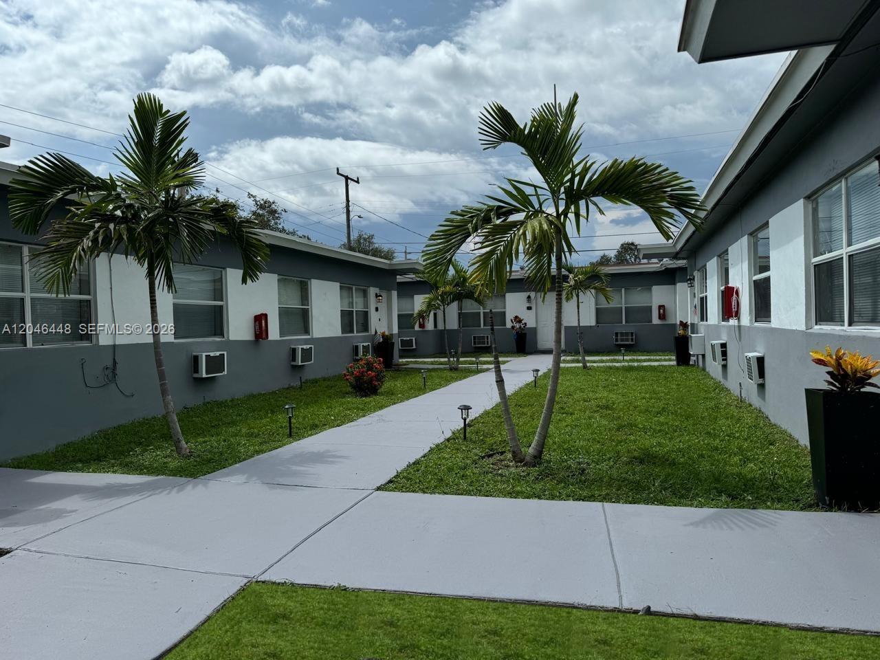 A paved walkway leads through a courtyard between two gray apartment buildings with palm trees — 2300 Douglas St # 1, Hollywood FL 33020, HOLLYWOOD, FL
