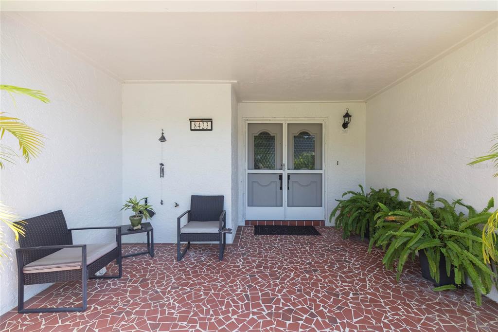 A covered front porch features a red mosaic tile floor, two chairs, and potted plants — 8423 PEBBLE DRIVE, PORT RICHEY, FL