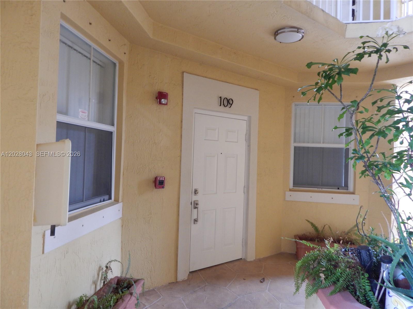 The exterior entrance to unit 109 features a white door, yellow stucco walls, and plants — 8964 W Flagler St # 109, Miami FL 33174, MIAMI, FL