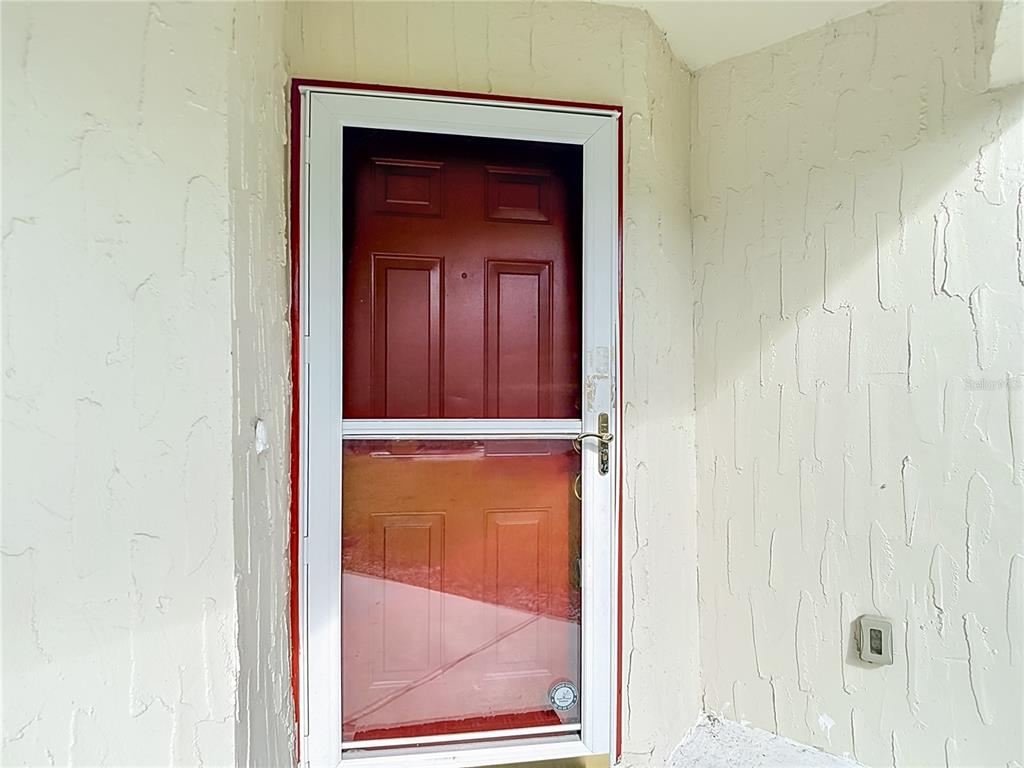 A red front door with a white storm door is set into a textured wall — 11543 ORLEANS LANE, PORT RICHEY, FL