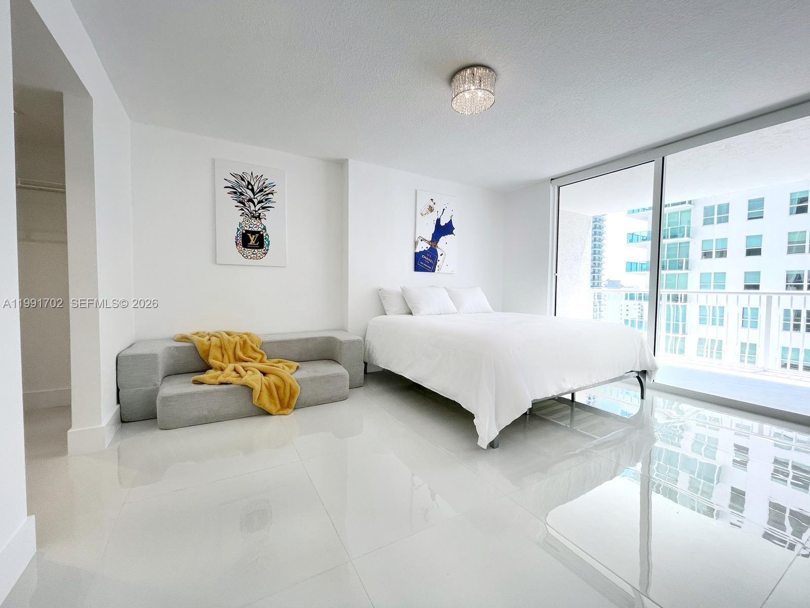 A modern bedroom features white tile floors, a bed, a low sofa, and balcony access — 1200 SE Brickell Bay Dr # 1707, Miami FL 33131, MIAMI, FL