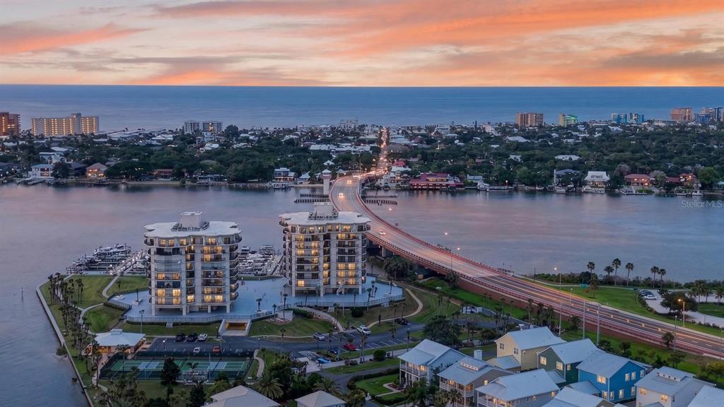 An aerial view shows two high-rise residential buildings, a bridge, and a coastal town at sunset — 503 N CAUSEWAY UNIT 2020, NEW SMYRNA BEACH, FL