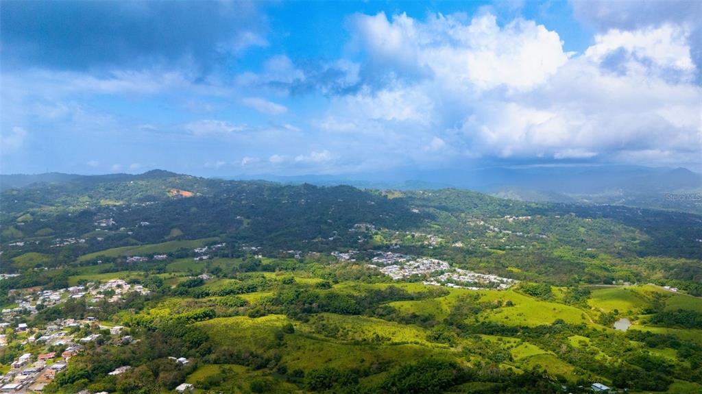 An aerial view shows a lush, hilly landscape with scattered houses and a cloudy sky — BO VALENCIANO ARRIBA CARR 919 KM 4.0, JUNCOS, PR