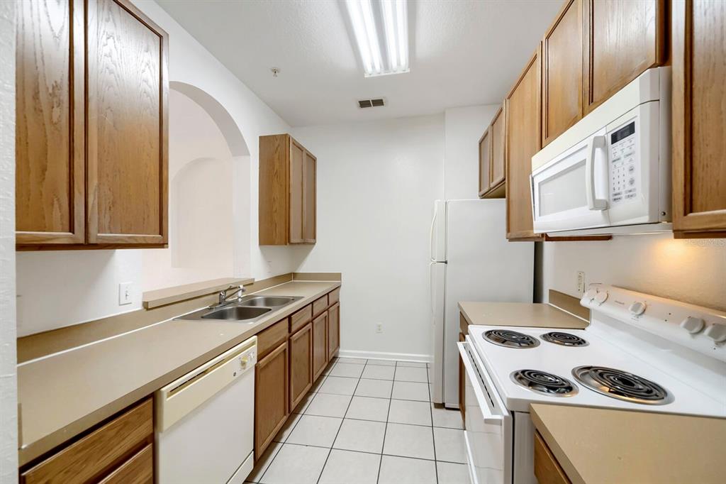A galley-style kitchen features wood cabinets, white appliances, laminate countertops, and white tile flooring — 6466 CAVA ALTA DRIVE UNIT 208, ORLANDO, FL