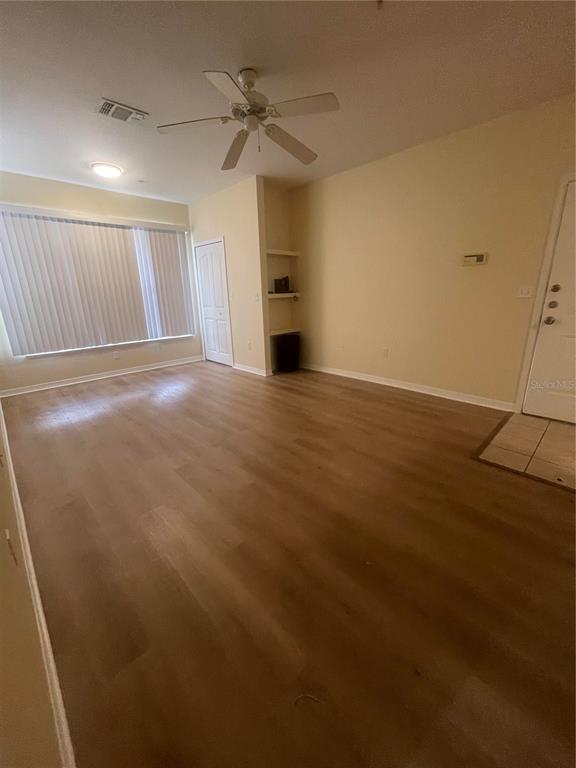 An empty living room features light wood-look flooring, a ceiling fan, and built-in wall shelving — 8820 DUNES COURT UNIT 202, KISSIMMEE, FL