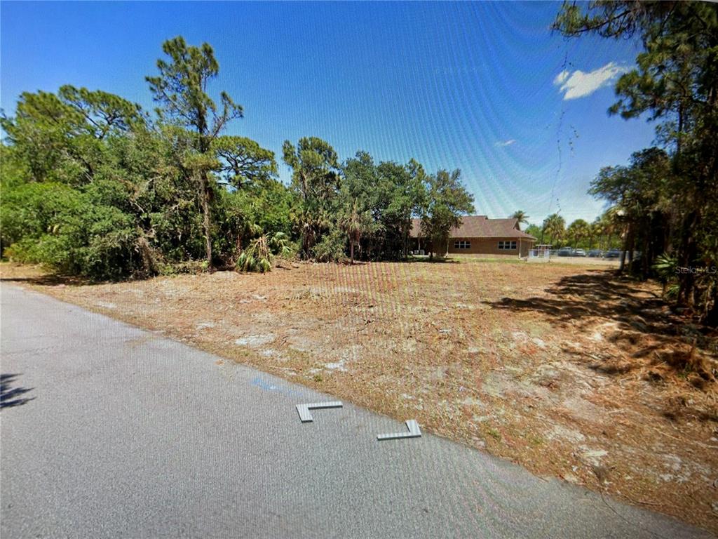 A vacant, grassy lot sits beside a paved road with trees and a house behind — 1412 LULLABY STREET, PORT CHARLOTTE, FL
