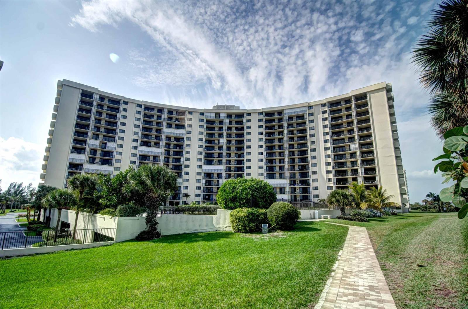 A large, curved multi-story apartment building stands behind a green lawn and paved walkway — 200 Ocean Trail Way # 109, Jupiter FL 33477, JUPITER, FL