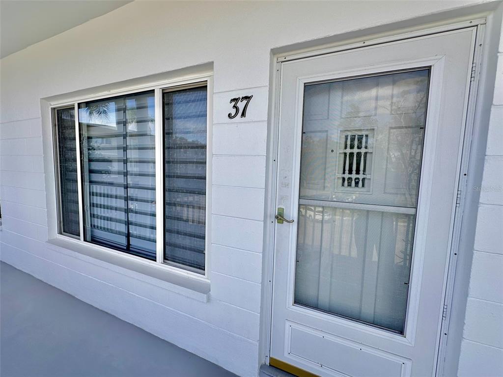 A white exterior wall features a window with horizontal blinds and a white storm door — 2431 CANADIAN WAY UNIT 37, CLEARWATER, FL