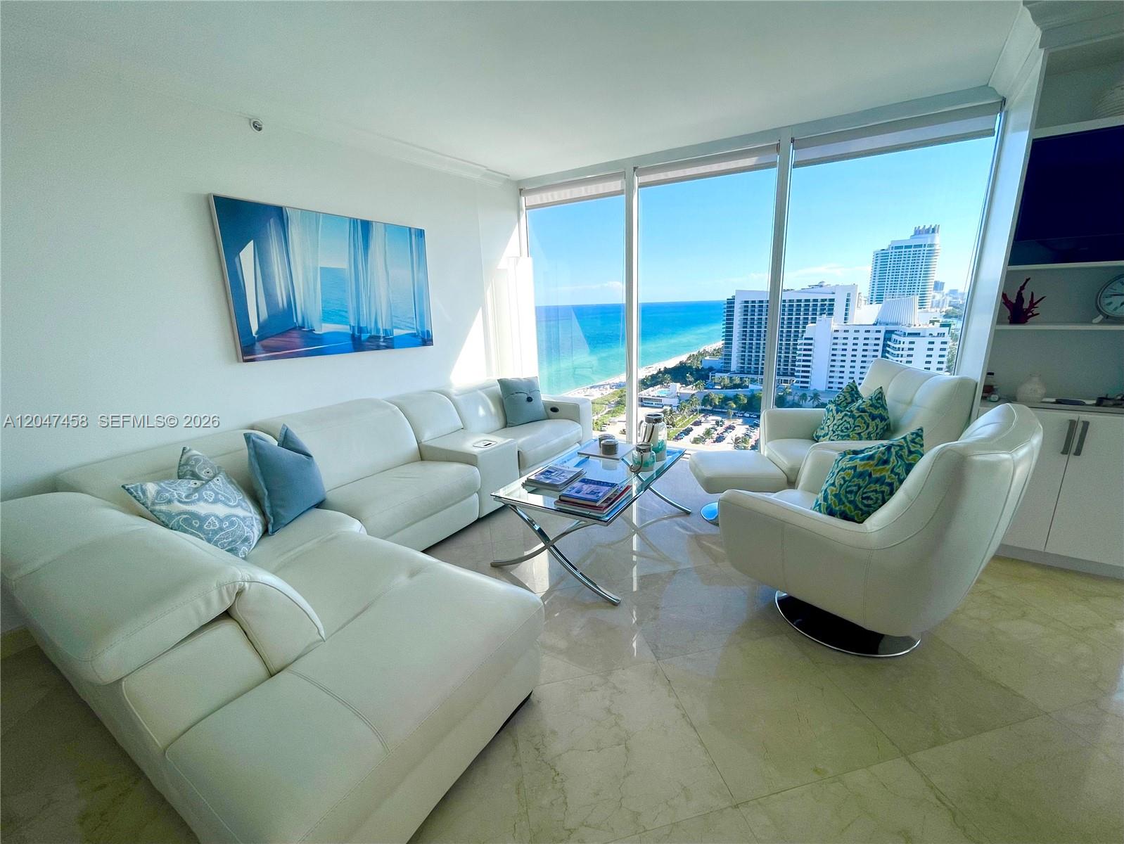 A bright living room features white leather furniture, a glass coffee table, and ocean views — 4775 Collins Ave # 2305, Miami Beach FL 33140, MIAMI BEACH, FL