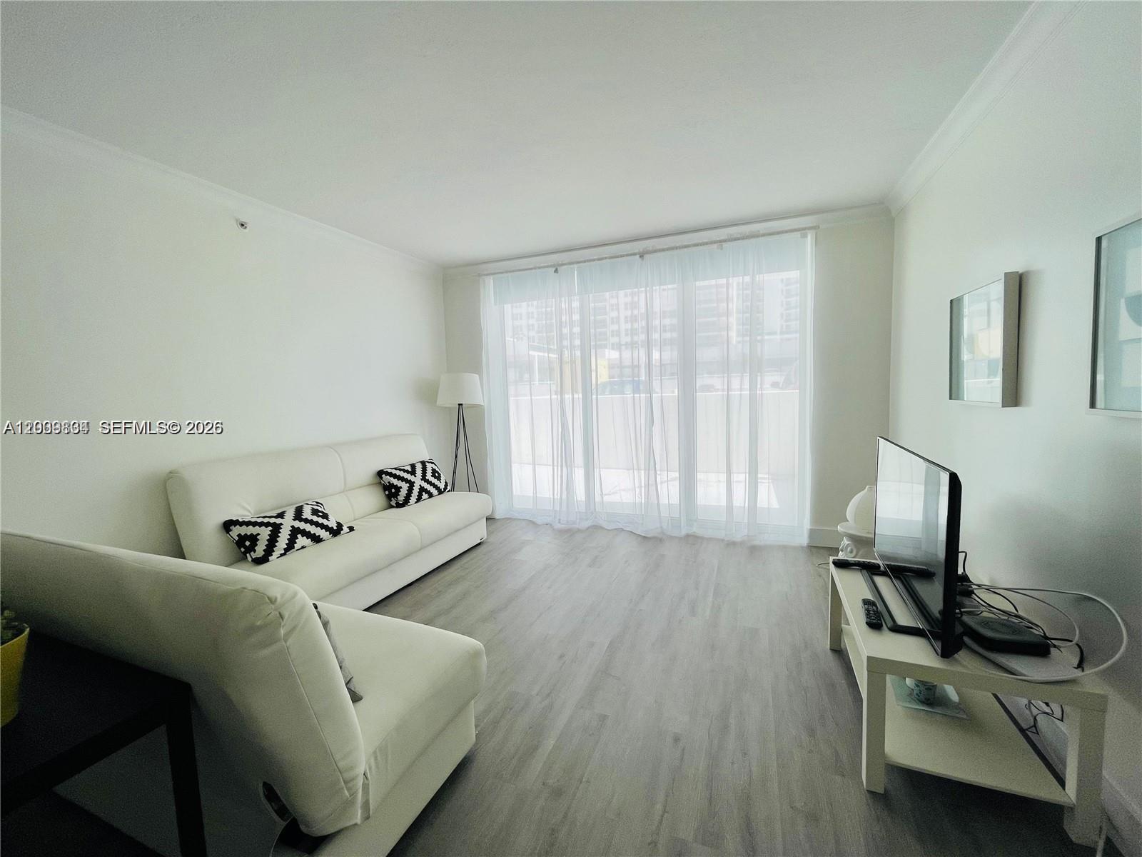 A living room features white sofas, light wood-look flooring, and a large window with curtains — 3001 S Ocean Dr # 101, Hollywood FL 33019, HOLLYWOOD, FL