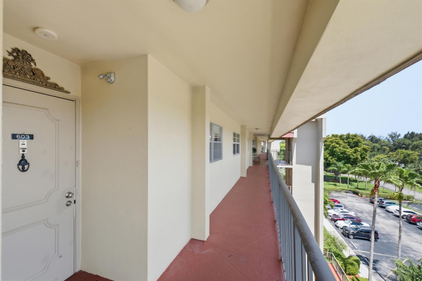 An outdoor apartment hallway with a red floor, metal railing, and a view of a parking lot — 3836 Via Poinciana # 603, FL 33467, FL