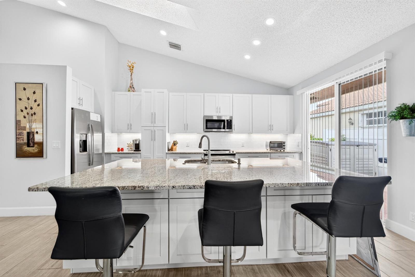 A modern kitchen features white cabinetry, a large granite island, and three black bar stools — 12902 Touchstone Place, Palm Beach Gardens FL 33418, PALM BEACH GARDENS, FL