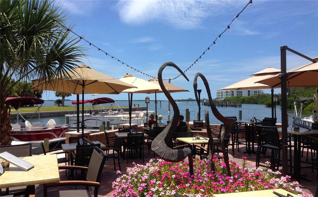 An outdoor waterfront patio features dining tables, umbrellas, and a metal sculpture among flowers — 12087 CLAREMONT DRIVE, PORT CHARLOTTE, FL