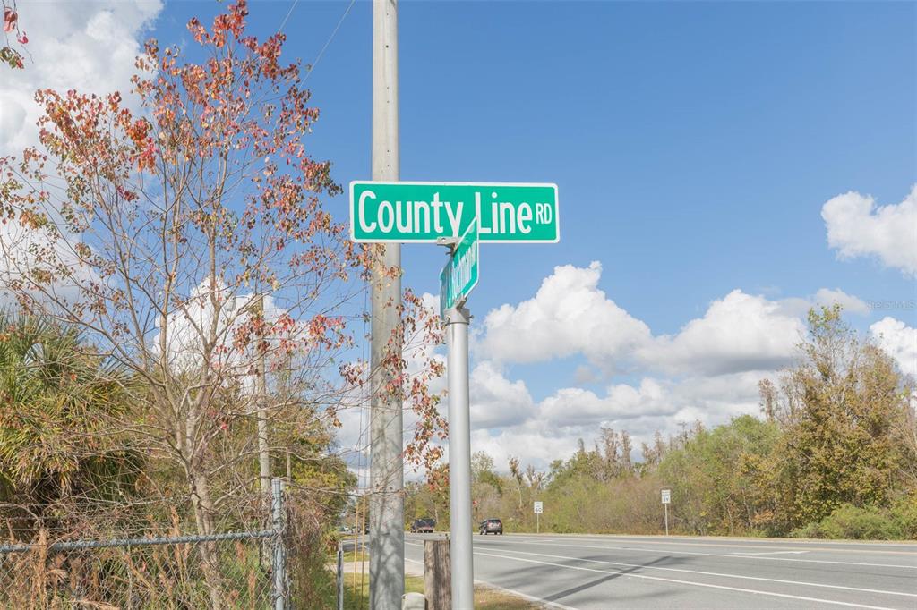 A street sign for County Line Road stands beside a road with trees and sky — 2050 PAUL S BUCHMAN HIGHWAY, ZEPHYRHILLS, FL