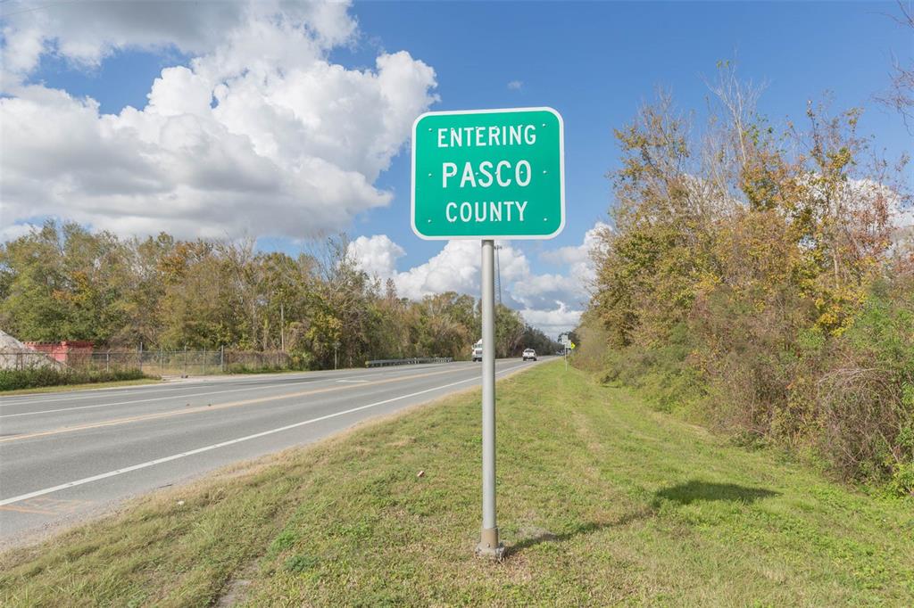 A green road sign reading Entering Pasco County stands beside a paved highway — 2050 PAUL S BUCHMAN HIGHWAY, ZEPHYRHILLS, FL