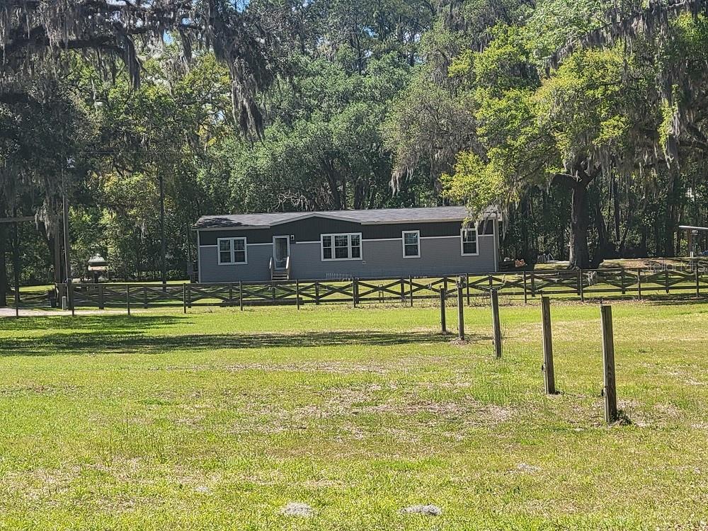 A single-story manufactured home sits behind a wooden fence in a grassy, tree-filled yard — 22397 MONDON HILL ROAD, BROOKSVILLE, FL