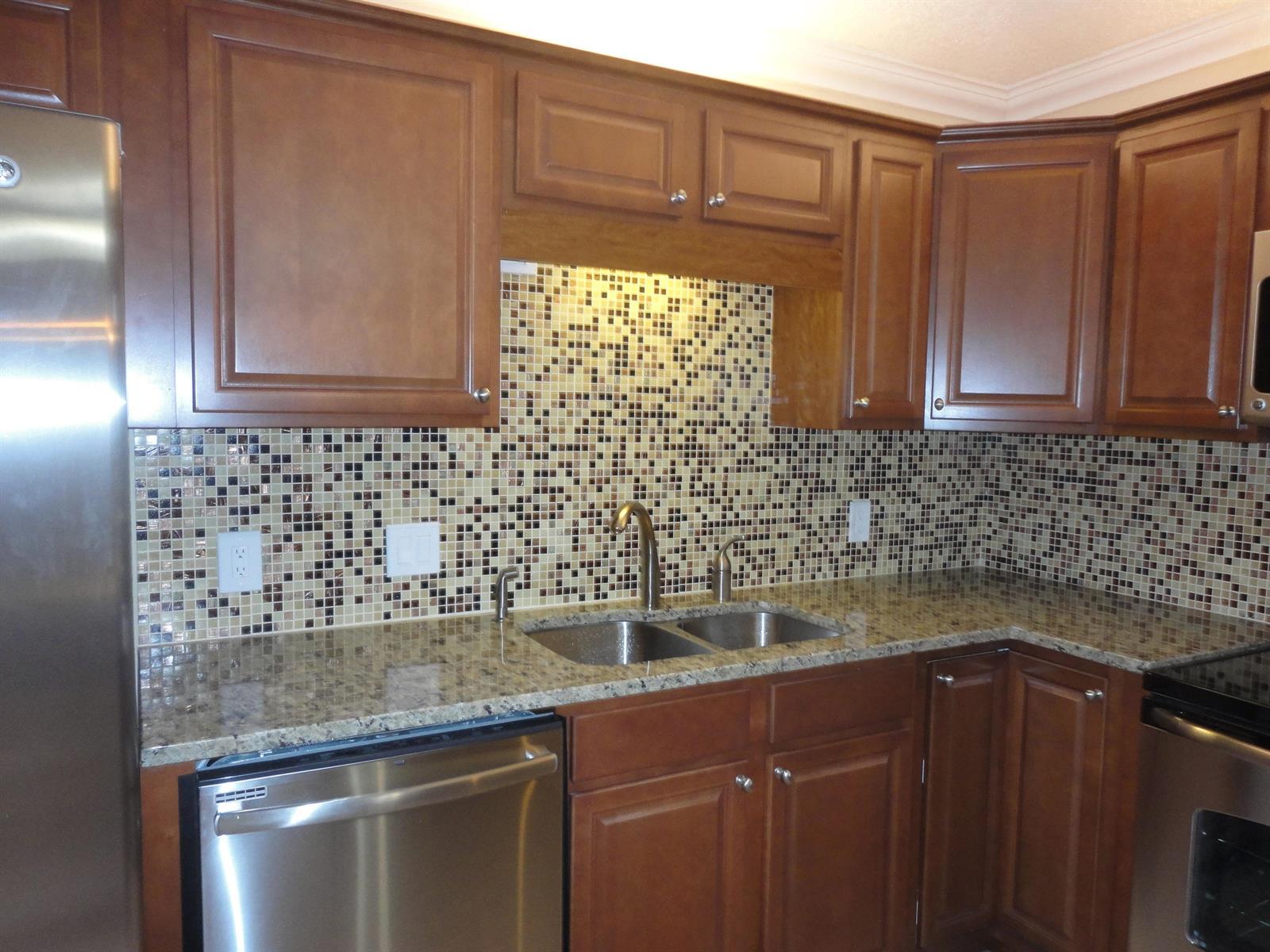 A kitchen features brown wood cabinets, granite countertops, a mosaic tile backsplash, and stainless appliances — 3008 San Clara Drive # b, Delray Beach FL 33445, DELRAY BEACH, FL