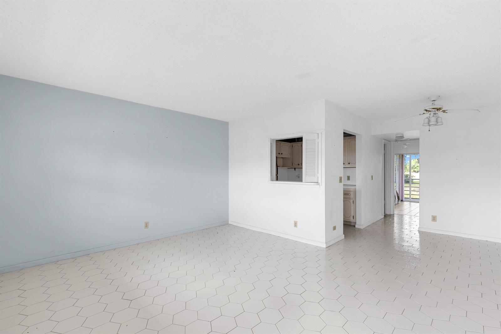 An empty room with light blue walls, white tile flooring, and a kitchen pass-through window — 72 Ashby B, Deerfield Beach FL 33442, DEERFIELD BEACH, FL