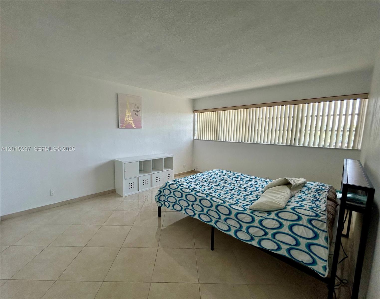 A bedroom features a bed with a patterned blue cover, tile flooring, and vertical blinds — 215 SE 3rd Ave # 408C, Hallandale Beach FL 33009, HALLANDALE BEACH, FL