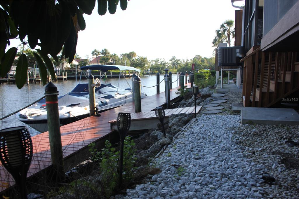 A wooden boat dock extends into a canal next to a gravel yard and house — 25188 MARION AVENUE UNIT 1027, PUNTA GORDA, FL