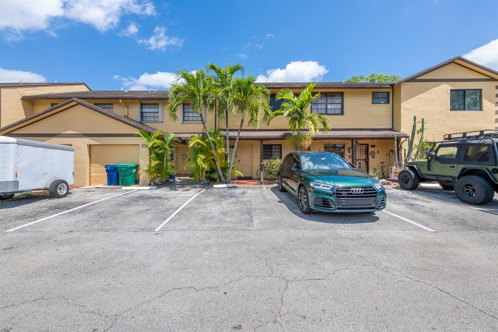 A two-story yellow townhouse exterior with a paved parking area and palm trees — 1129 NE 210th Terrace # 210, Miami FL 33179, MIAMI, FL