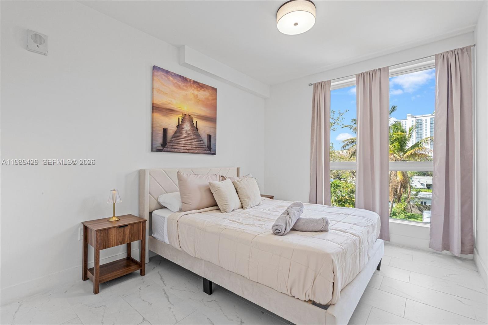 A bedroom features a bed with a beige headboard, a wooden nightstand, and large windows — 1850 Monroe Street # 309, Hollywood FL 33020, HOLLYWOOD, FL