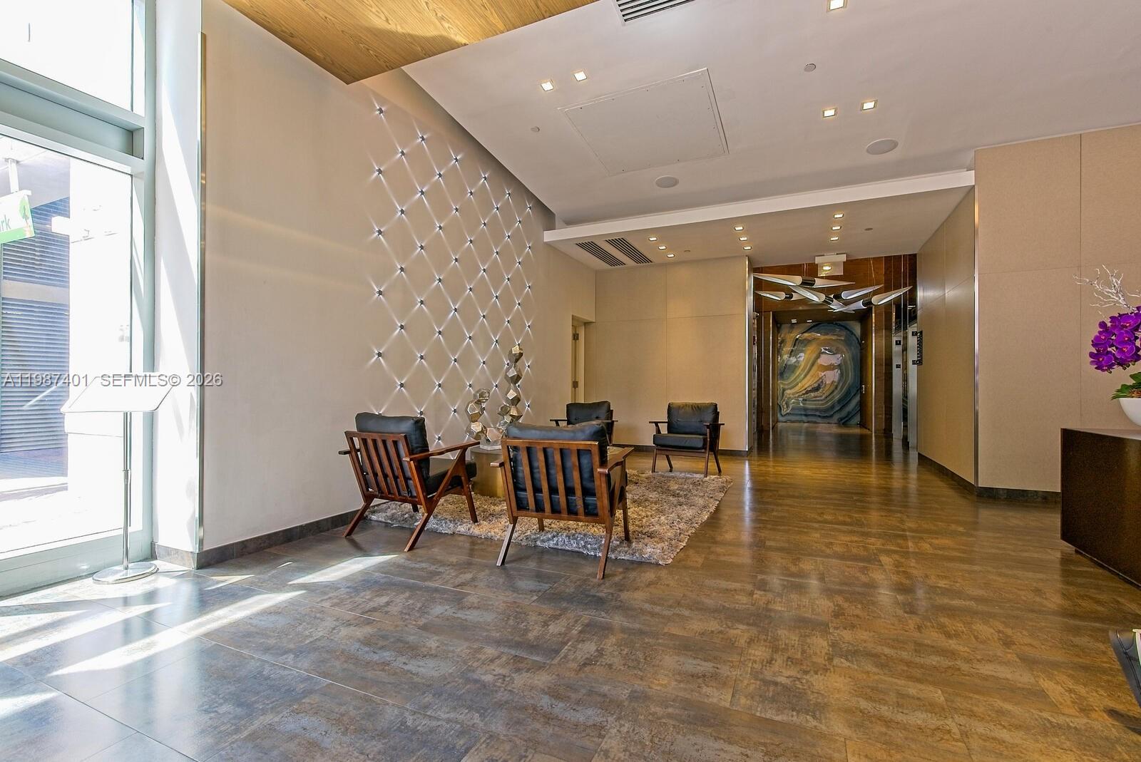 A modern building lobby features dark wood-framed chairs, a textured wall, and tile flooring — 3250 NE 1st Ave # 1006, Miami FL 33137, MIAMI, FL