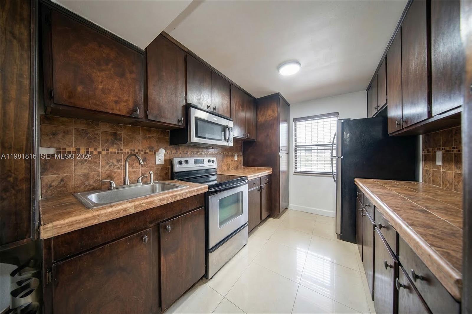 A galley kitchen features dark wood cabinets, brown tile countertops, and stainless steel appliances — 10710 NW 7th St # 11B, Miami FL 33172, MIAMI, FL