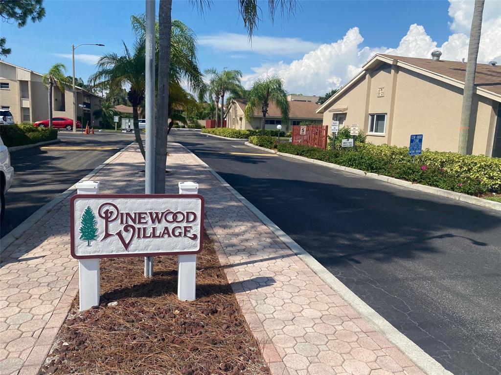 A Pinewood Village sign stands on a paved walkway in front of apartment buildings — 2763 WOODGATE LANE UNIT 208, SARASOTA, FL
