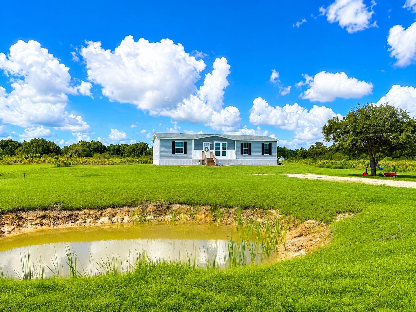 A light blue manufactured home sits on a grassy lot behind a small pond — 18720 NW 242nd Street, Okeechobee FL 34972, OKEECHOBEE, FL