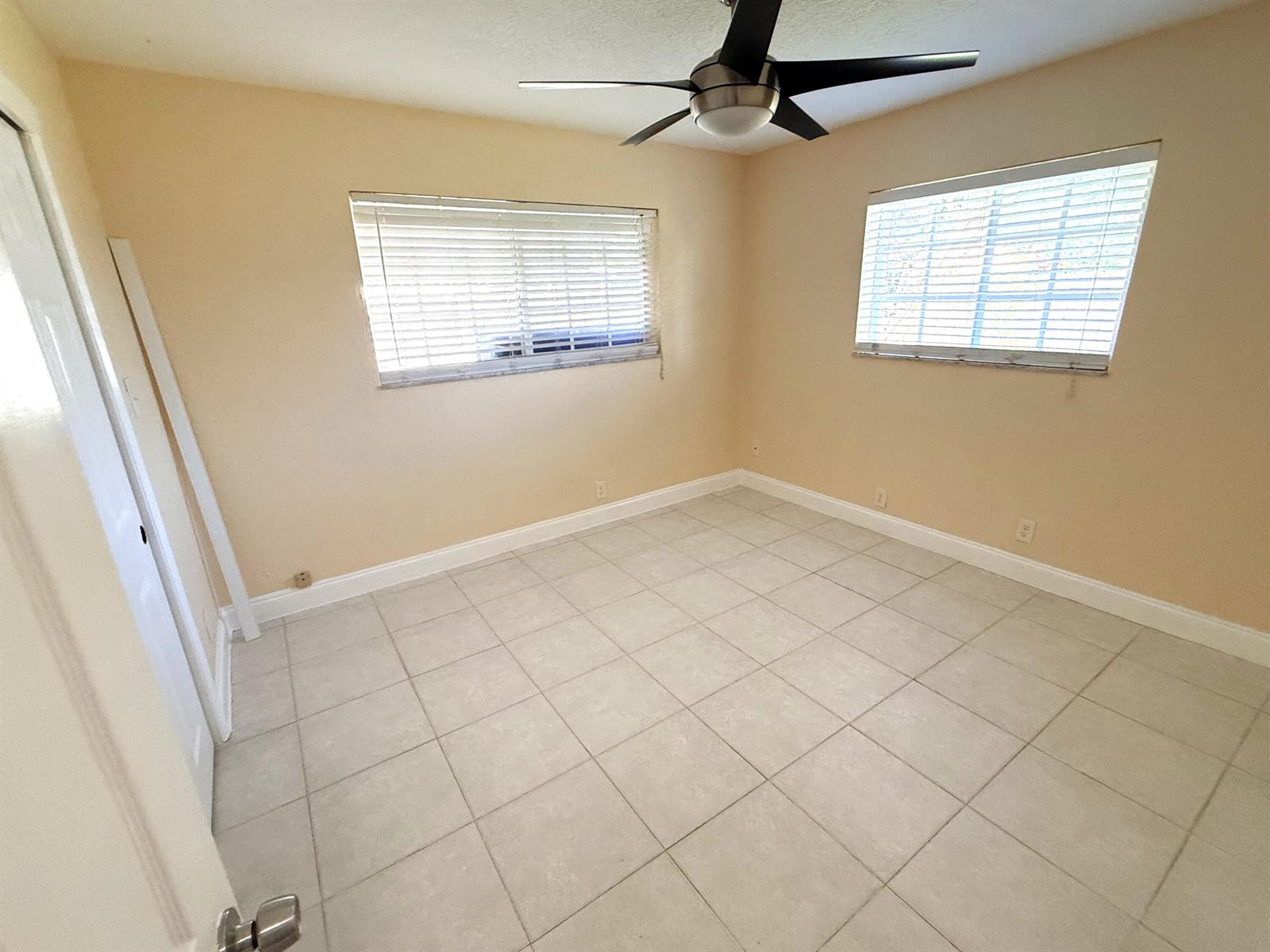 An empty bedroom features beige walls, light-colored tile flooring, two windows with blinds, and a fan — 551 SE 15th Street, Pompano Beach FL 33062, POMPANO BEACH, FL