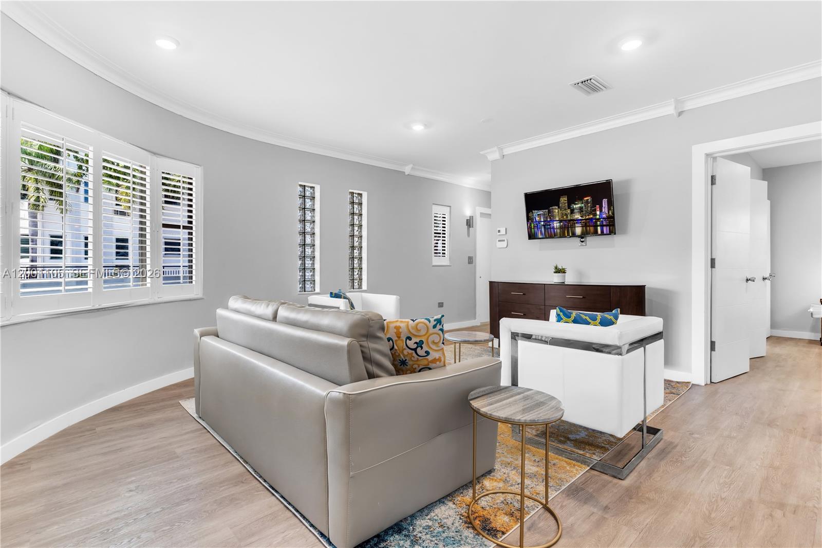 A living room features a gray sofa, white armchairs, wood-look flooring, and a wall-mounted television — 8101 Harding Ave, Miami Beach FL 33141, MIAMI BEACH, FL