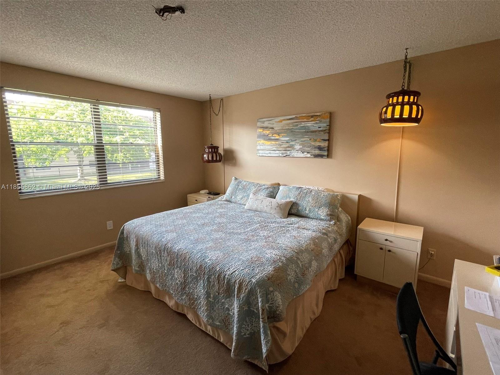 A bedroom features a bed with a patterned quilt, two hanging pendant lights, and carpeted flooring — 4975 E Sabal Palm Blvd # 208, Tamarac FL 33319, TAMARAC, FL