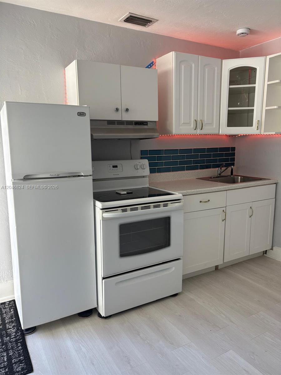 A small kitchen features white cabinets, a white stove, a refrigerator, and blue backsplash tiles — 1101 NE 5th St # 1, Hallandale Beach FL 33009, HALLANDALE BEACH, FL