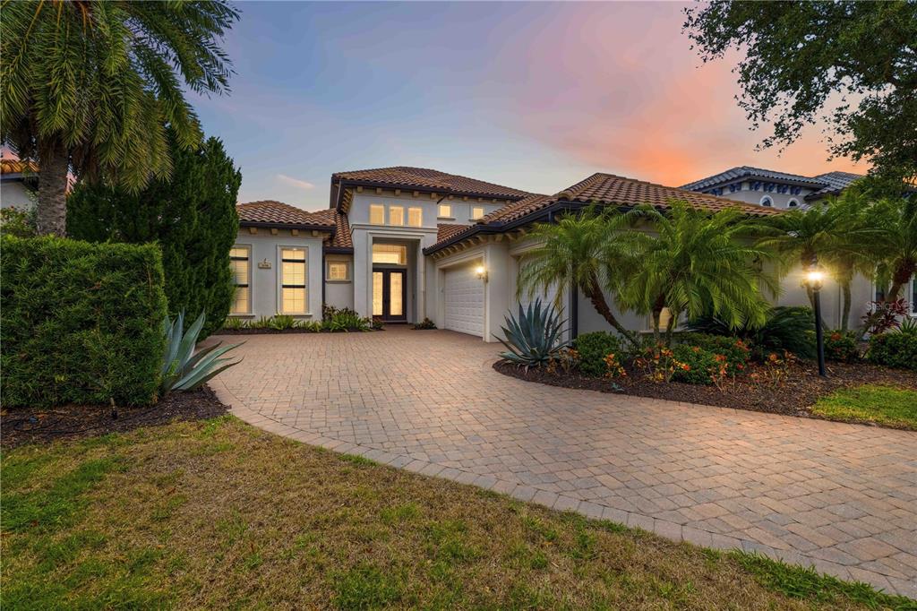 A single-story house with a tiled roof, paved driveway, and palm trees at sunset — 7638 WINDY HILL COVE, BRADENTON, FL